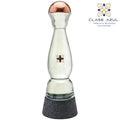 A clear glass tequila decanter with a copper-colored top, a small black floral emblem on the front, and a textured dark gray base. The Clase Azul logo is visible in the upper right corner against a plain white background.